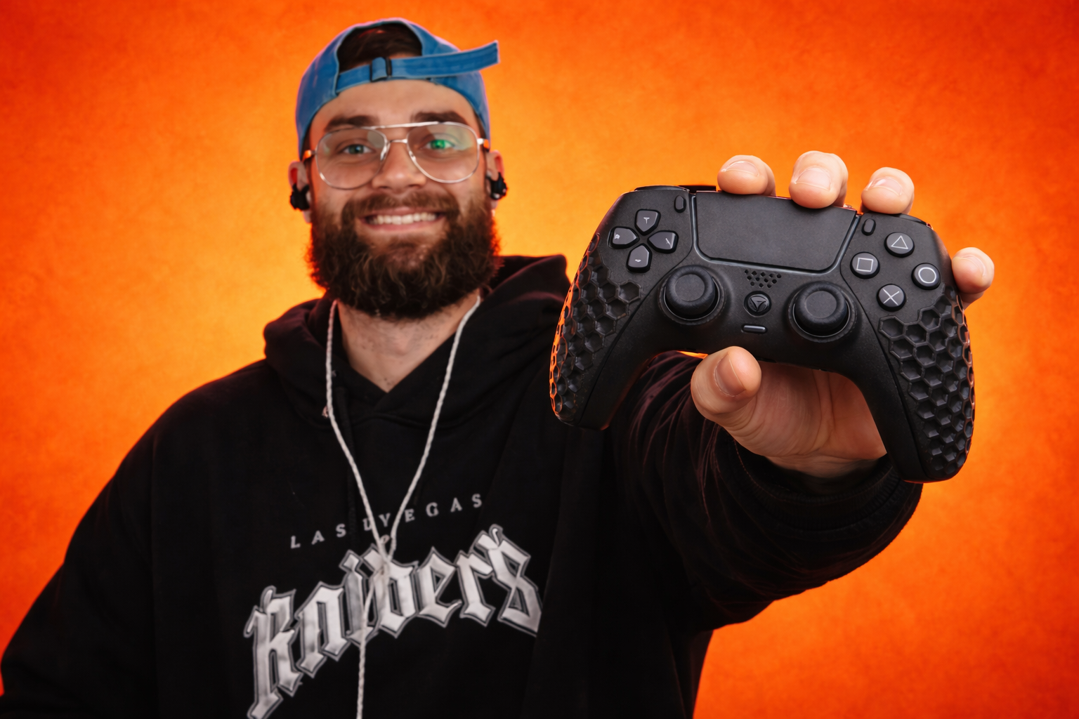 A smiling man with glasses and a beard holding out a black Helico Hexavent gaming controller towards the camera against a solid vibrant orange background.