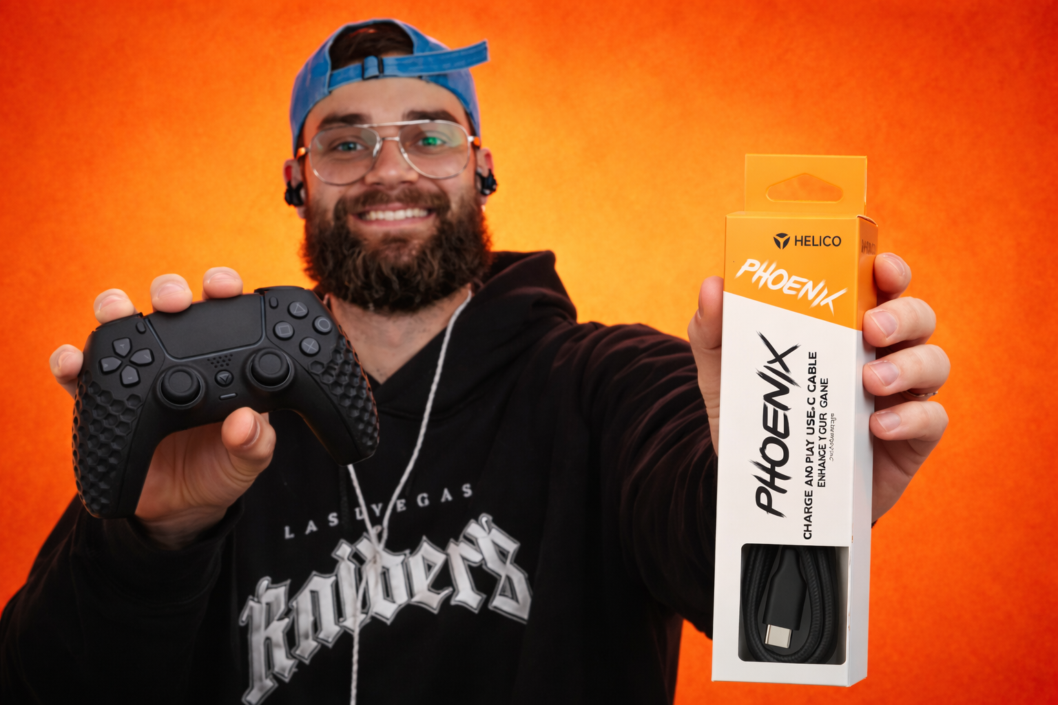 A smiling man holding a black Helico Hexavent gaming controller in one hand and a boxed Helico Phoenix USB-C Charge and Play cable in the other against a bright orange background.